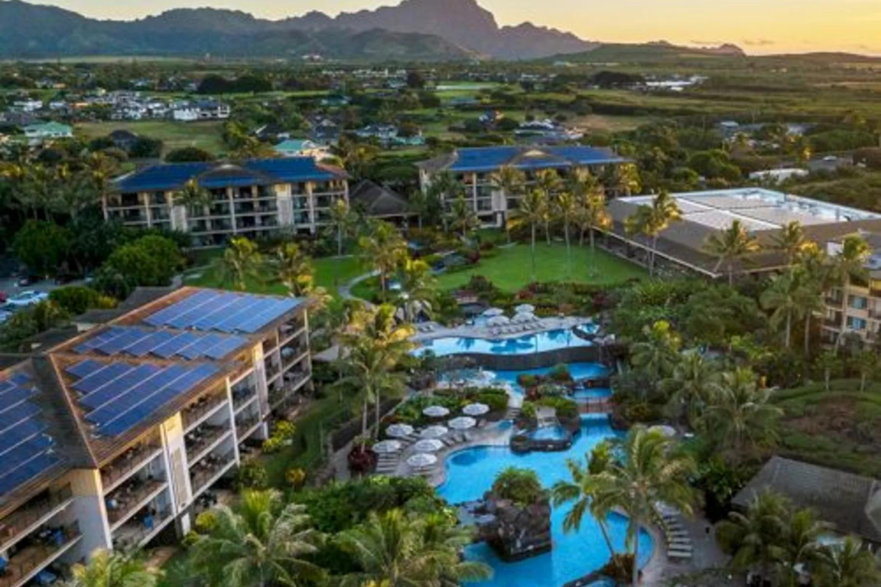 Aerial view of a resort with pools, palm trees, and buildings, surrounded by lush greenery and mountains.
