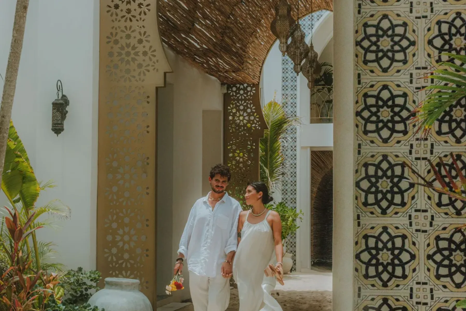 Couple walking through ornate archway, holding hands, in a courtyard with plants and decorative tile.