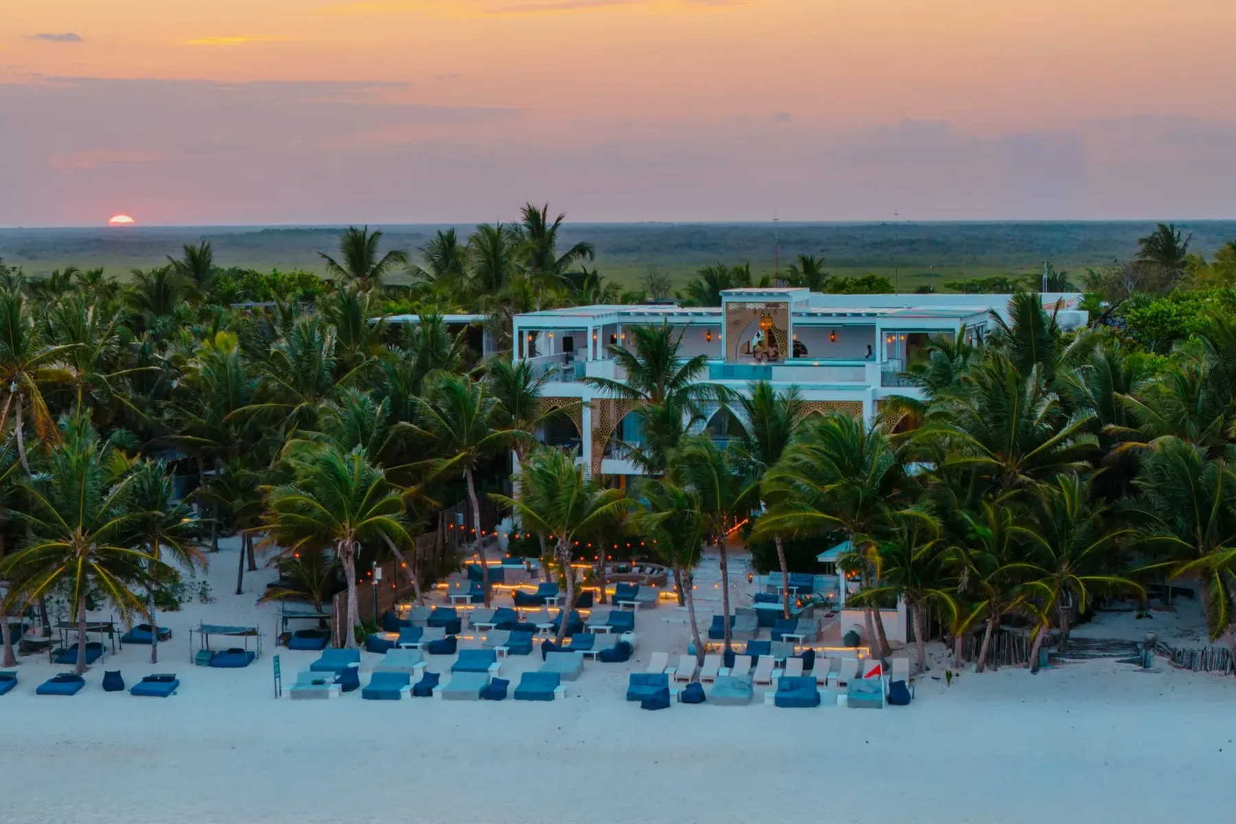 A beach resort with a white building nestled among palm trees at sunset, featuring lounge chairs on the sand.